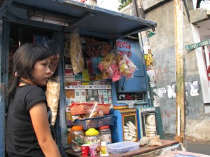 A girl in front of a street corner stall in Jakarta selling cigarettes, candy, and crackers.  There are tens of thousands, if not more, of such street stalls in Jakarta.                         